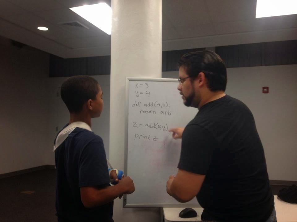 Picture of two people in front of a whiteboard in a white room. The older of the two is explaining some computer programming concepts.